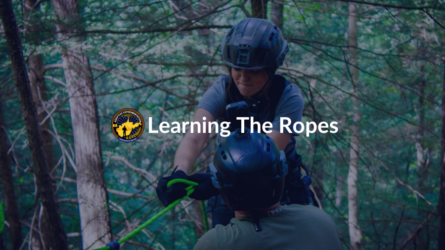 An individual in a helmet and safety harness uses ropes for rappelling as an instructor watches.