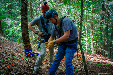 An individual in a helmet and safety harness uses ropes for rappelling as an instructor watches. 